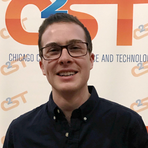 A photo of James Schwabacher smiling, wearing glasses, a black Oxford button-up shirt, standing in front of a C2ST background 